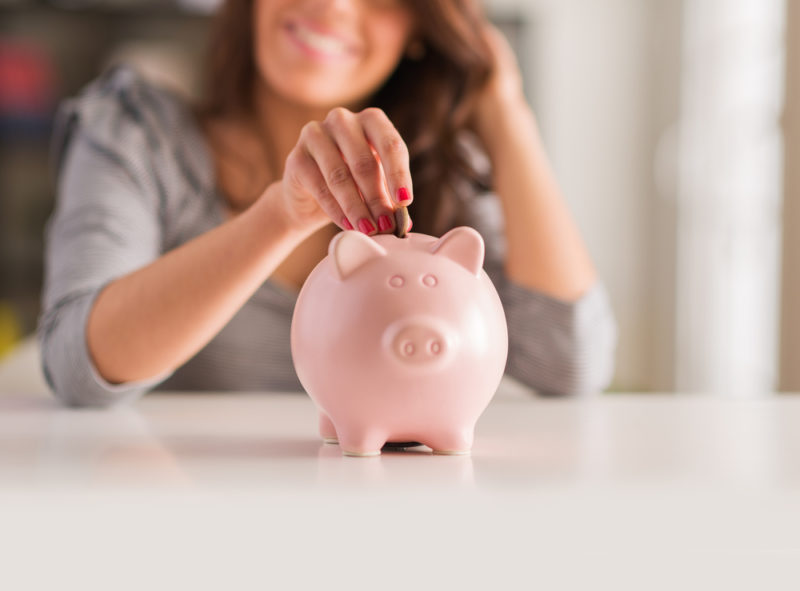 Porcelain piggy bank on a table; woman in the background is out of focus as she slips a coin into the top.
